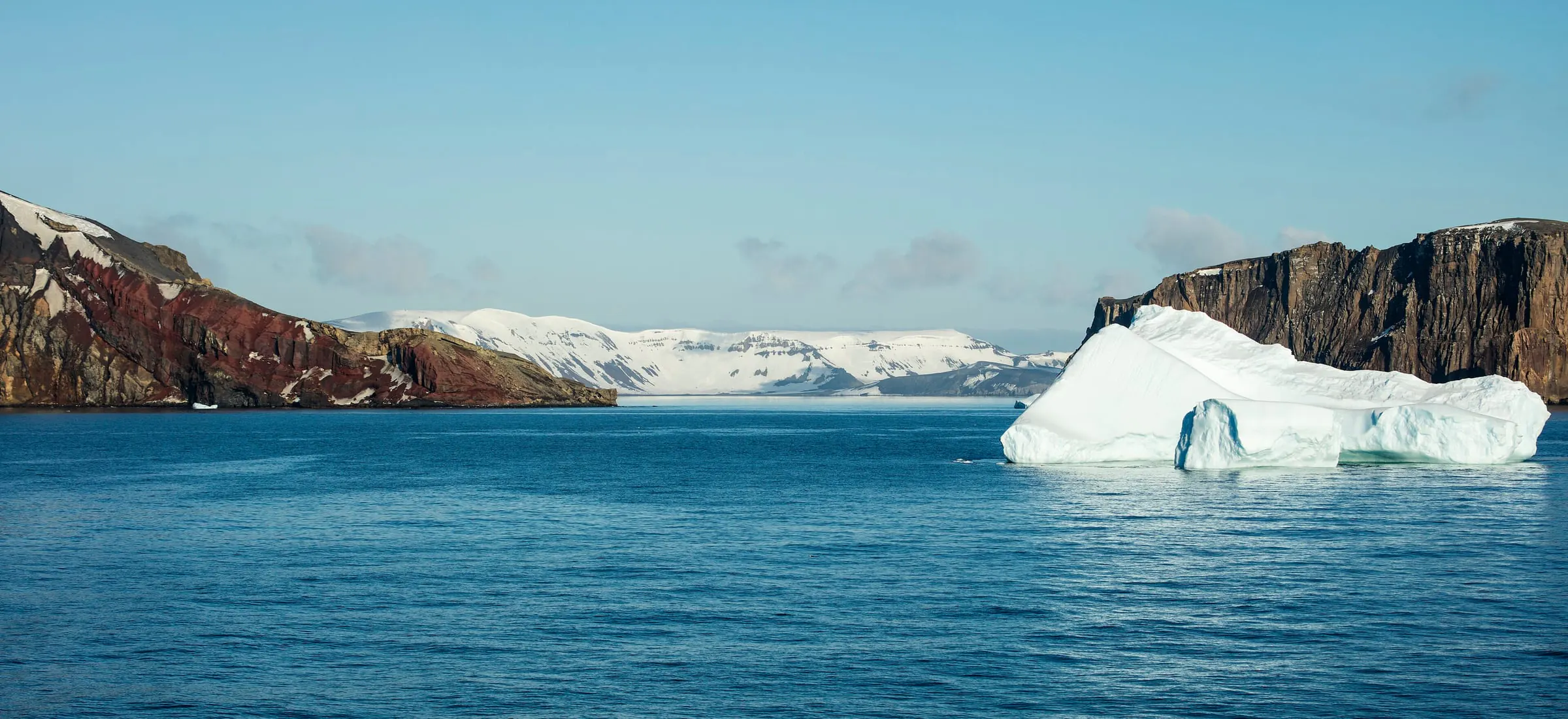Dark volcanic cliffs frame the narrow entrance of Neptune’s Bellows at Deception Island, with an iceberg floating on blue water.