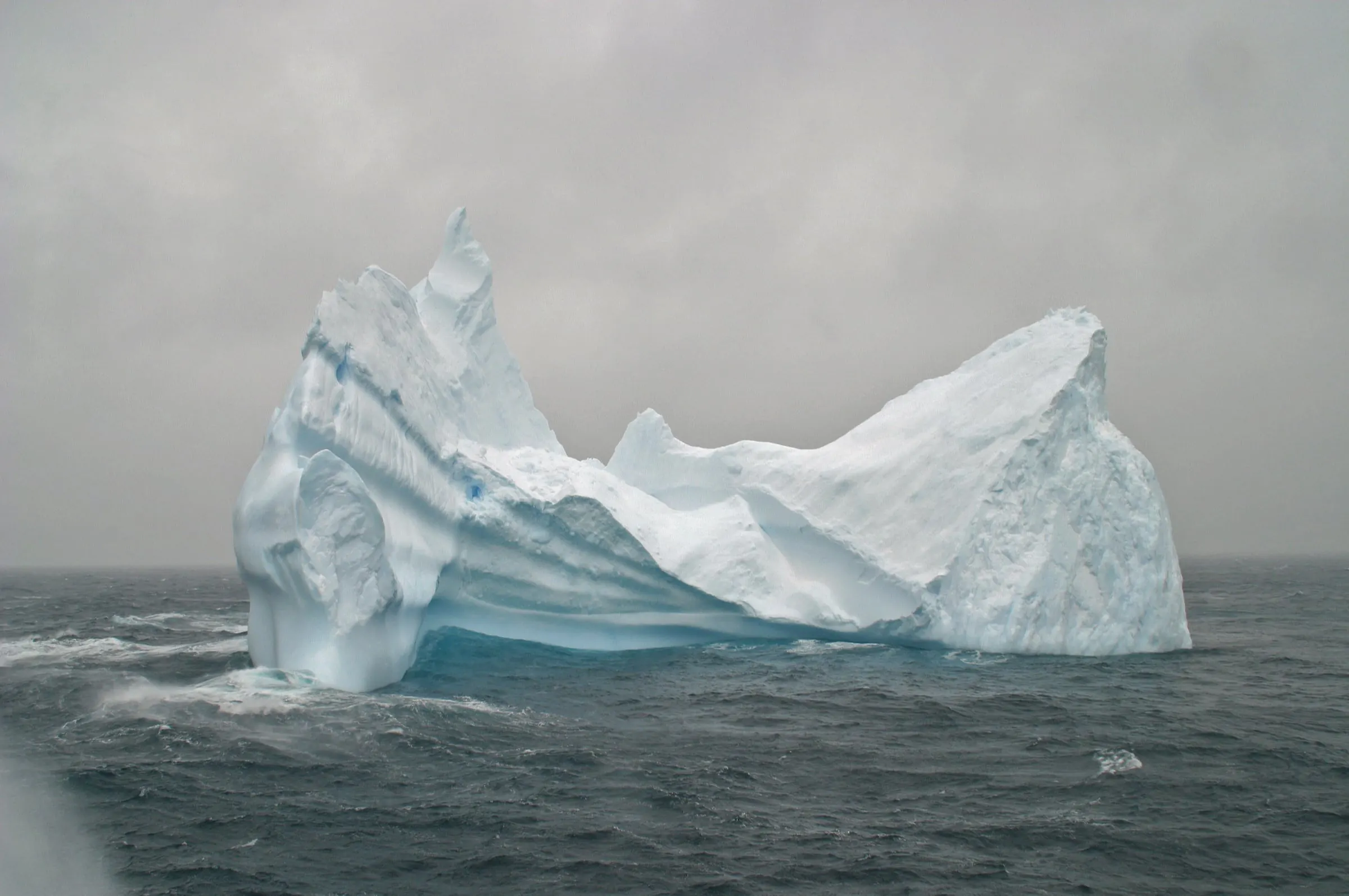 A large sculpted iceberg drifts in the rough grey waters of the Drake Passage under a stormy sky.