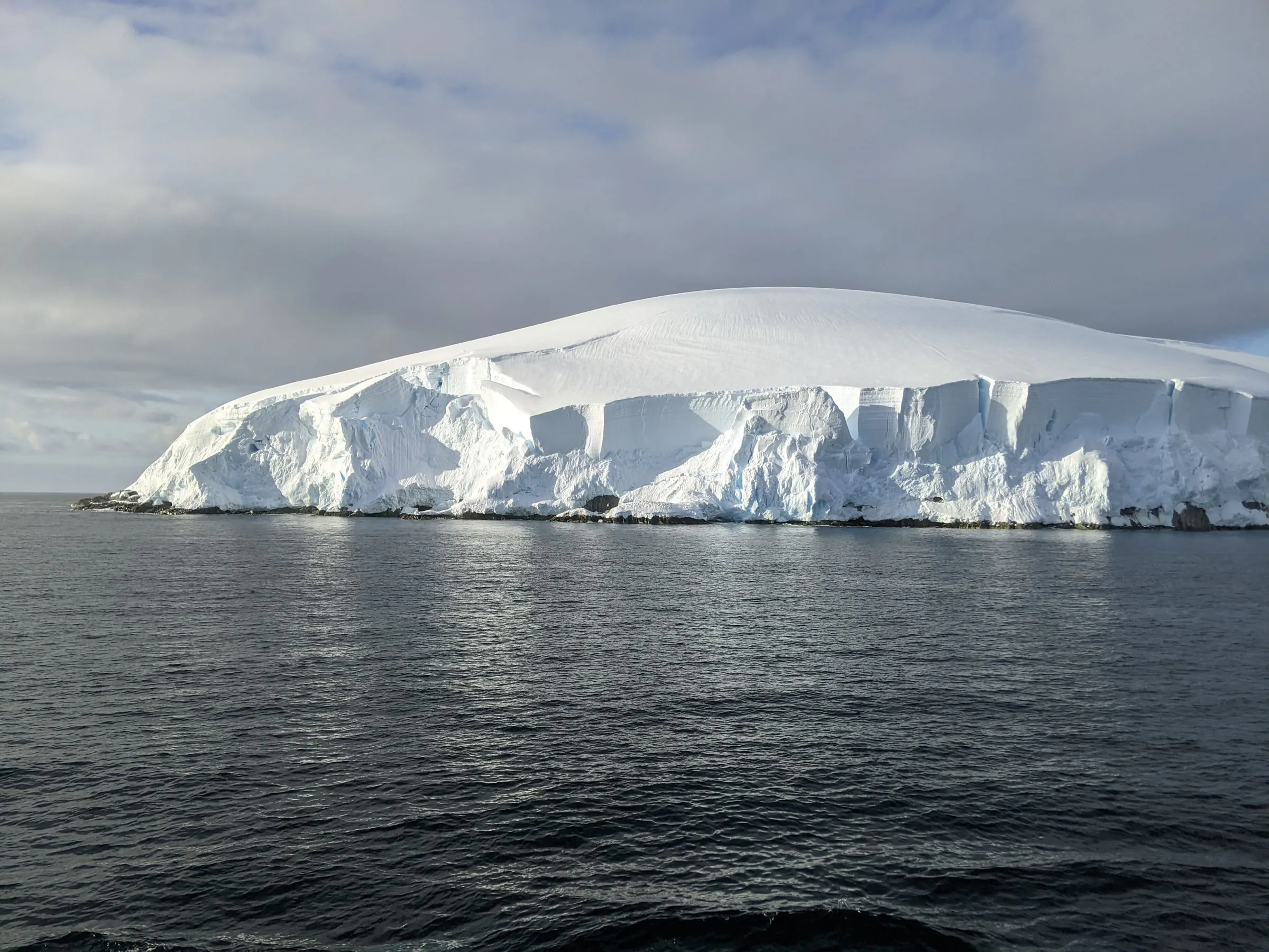 A snow-domed island with ice cliffs rises from calm dark water in the Melchior Islands.