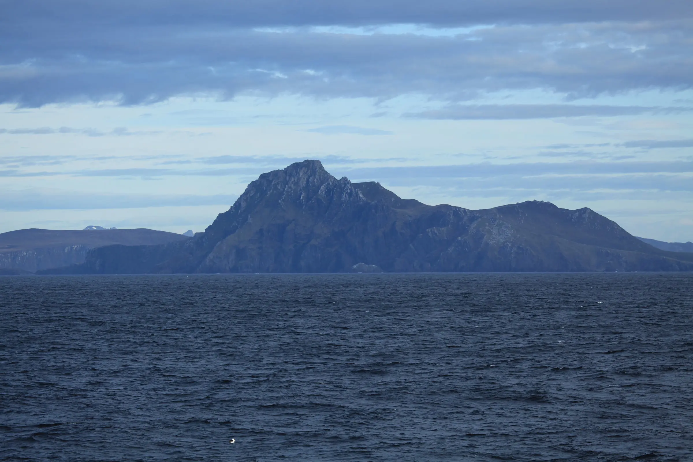 The dark rocky headland of Cape Horn rises from the Southern Ocean under a soft blue evening sky.