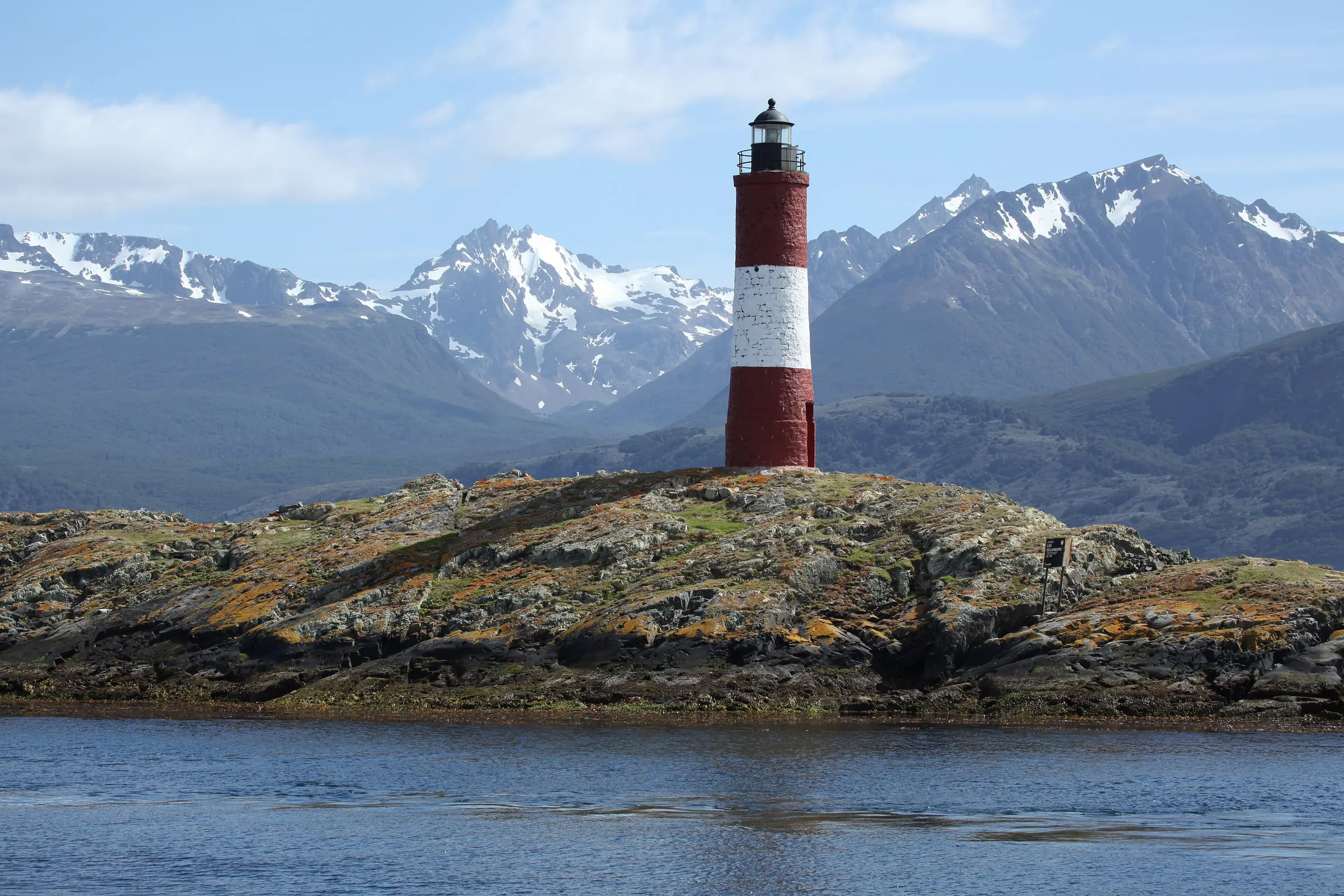 The red and white Les Eclaireurs lighthouse stands on a rocky islet in the Beagle Channel with snow-capped mountains behind.
